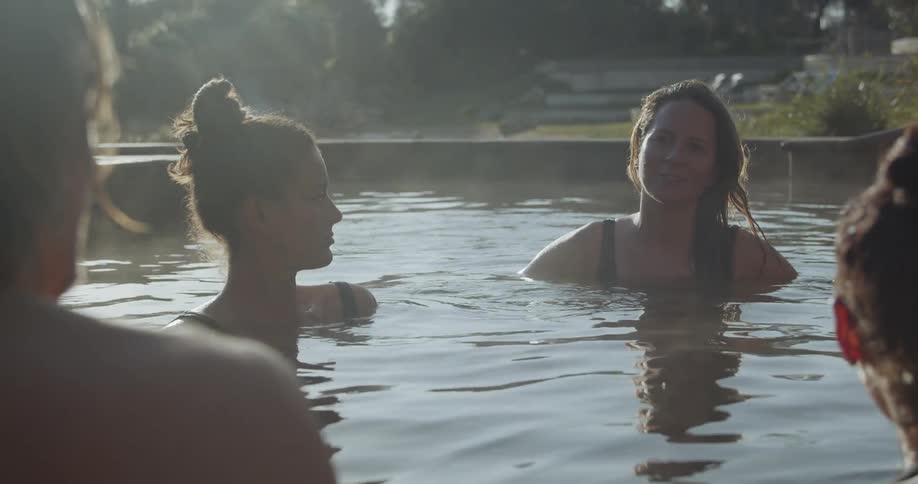 A group of people sit and converse in a large outdoor hot tub as steam rises from the surface