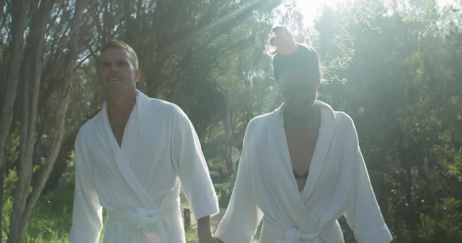 A couple wearing white bathrobes smile and hold hands as they walk outdoors on a sunny day