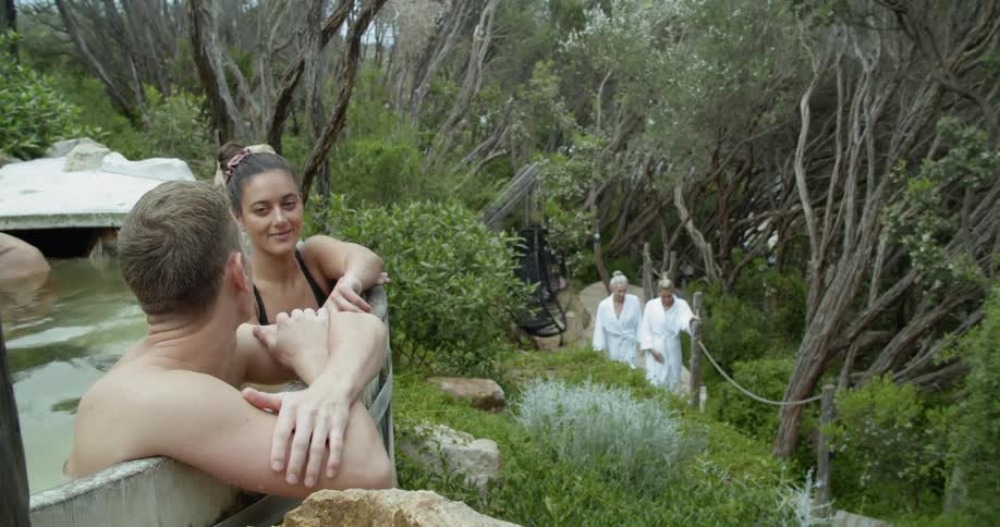 People relaxing in a kind of thermal bath surrounded by bushes as a couple of women walk up a path to join them