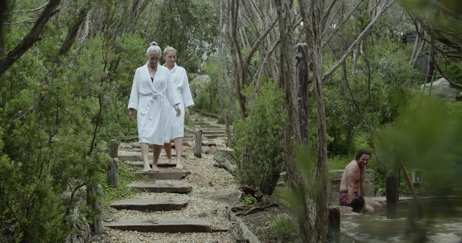 Two women in robes walk along a path past a hot tub