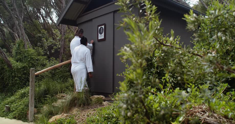 Two people in white terry cloth robes slide open the door of an outdoor sauna