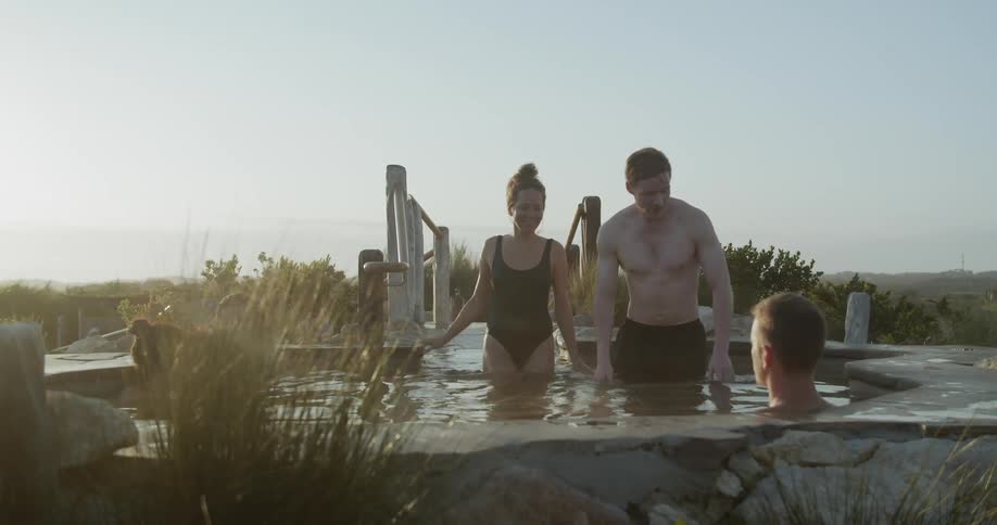 A couple smiles as they enter a large outdoor spa and join the others who are already relaxing in it
