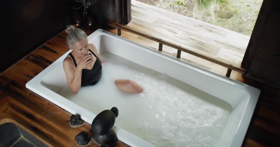An elderly woman in a bathing suit sits in a tub and drinks out of a cup