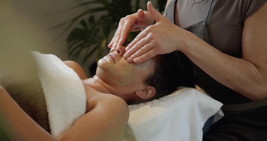 A female masseuse sits at a massage table where she focuses on giving a facial massage to the woman laying on the table