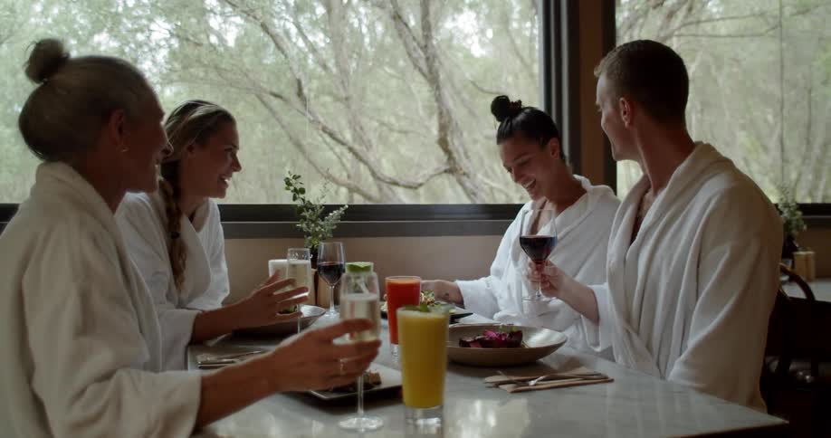 Four people in white robes are enjoying a meal and glasses of wine in a restaurant with large windows