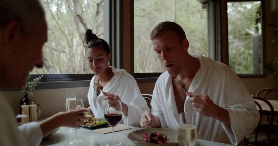 Four people dressed in white spa robes are sitting at a table in a restaurant eating a meal and drinking wine