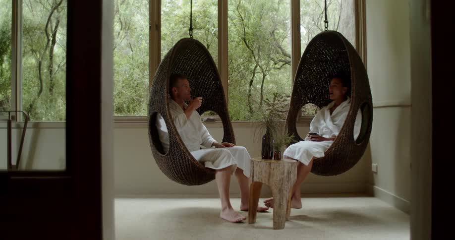 A couple wearing robes sit in hanging chairs and drink a beverage