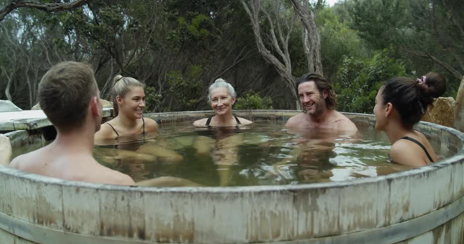 Six white people are soaking in a large outdoor hot tub with dense trees behind them
