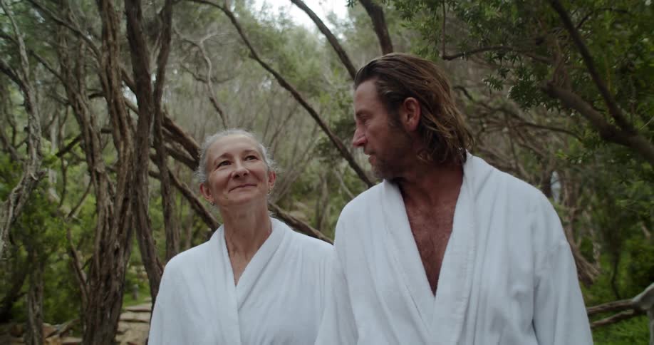 A man and woman wearing white bathrobes talk while they walk outside on a path through many trees