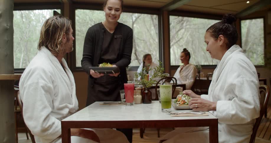 A man and woman wearing white robes are seated in a dining area as a server places their entrees on the table they are seated at