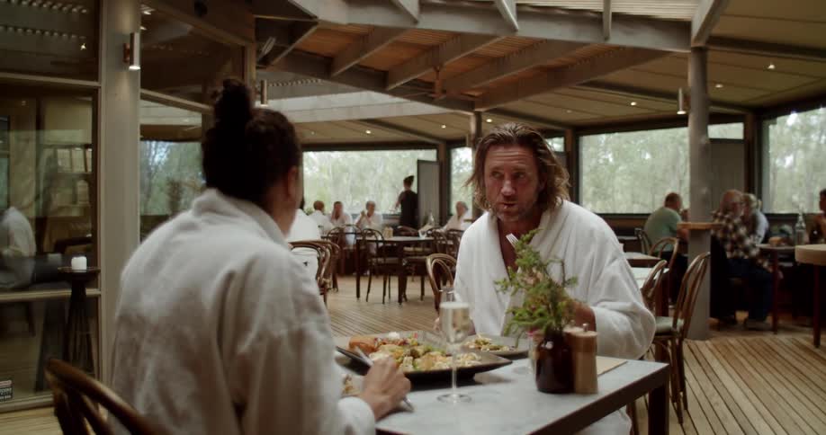 A man and woman sit at a table and eat in a dining area while wearing white bathrobes
