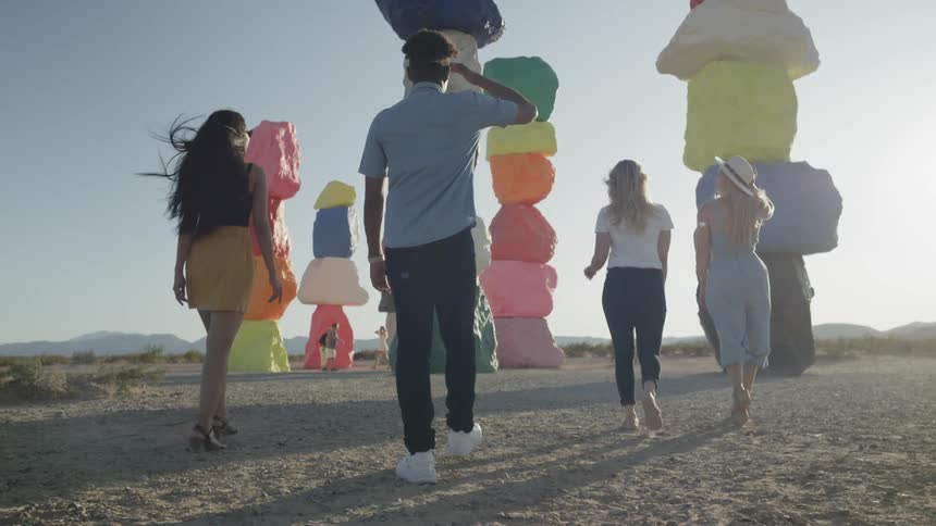 A group of young people are walking outdoors towards multiple columns of stacked boulders painted with bright colors