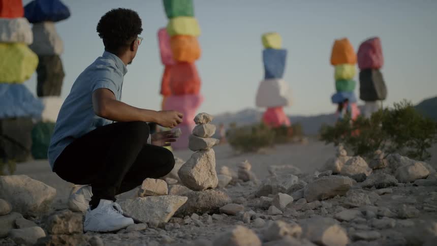 A young black male is crouching down and stacking some small stones at several larger colorful pillars of stones can be seen in the background