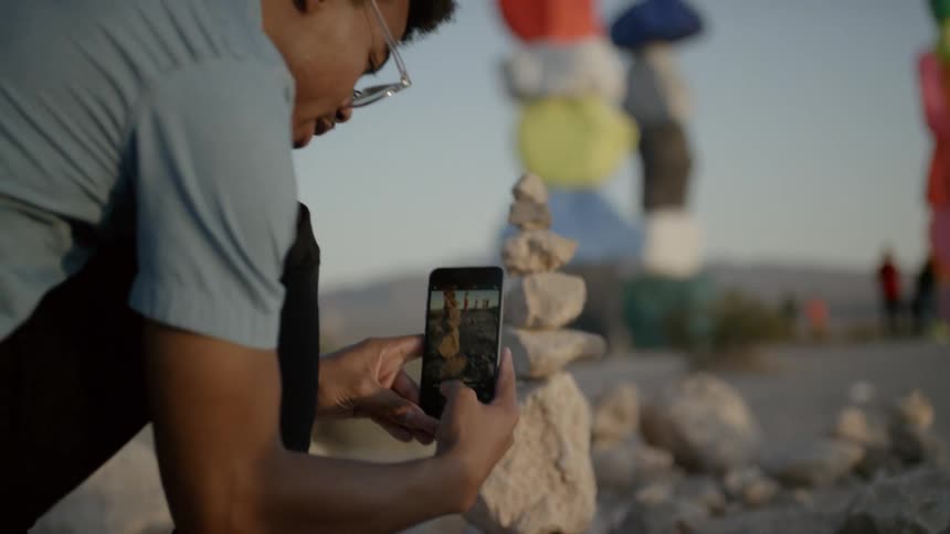 A young man kneeling to take photos of a stack of stones with his smartphone in the desert