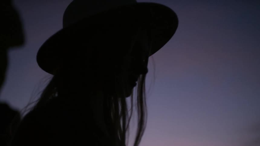 A woman is standing outdoors wearing a hat and watching the sun set