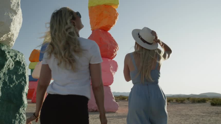 Two blonde women walk between colorful stone pillars outdoors on a windy day