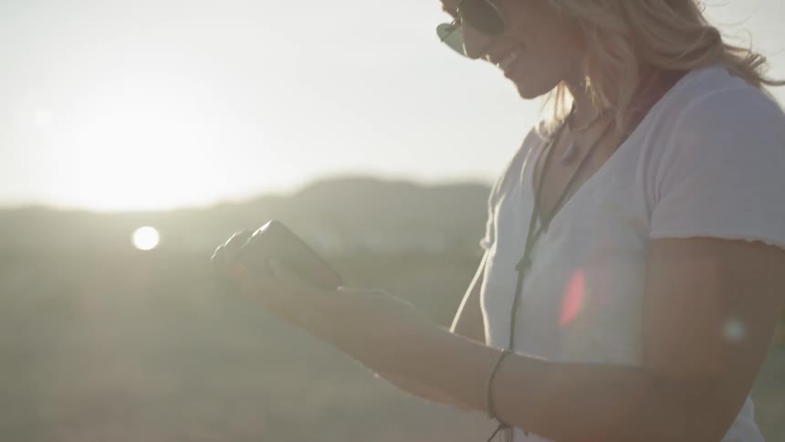 A blonde haired woman wearing sunglasses plays with a smart phone at low sun