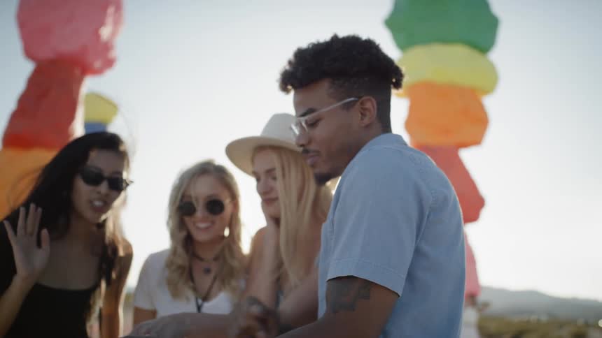 A group of friends gathered around a phone laugh and talk near colorful painted rocks in the desert