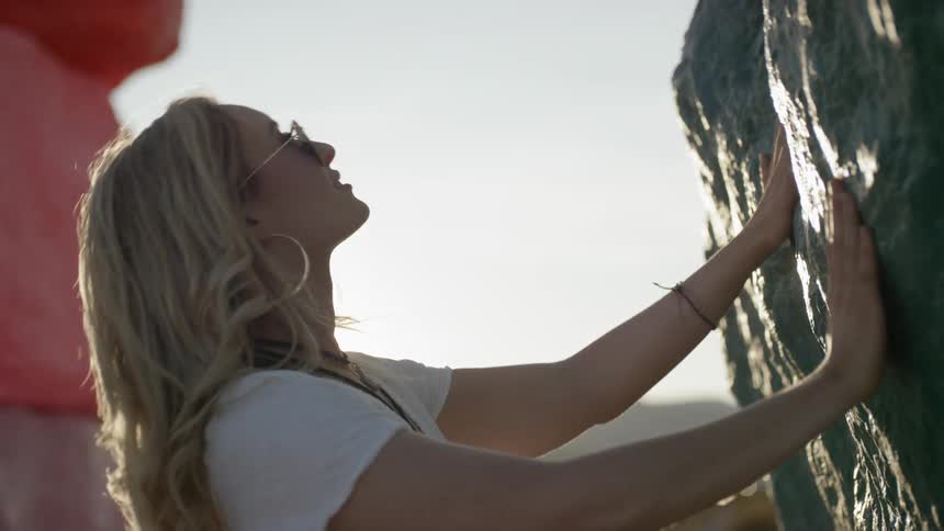 A woman touches rock with both hands and leans back to look up as the sun shines in the desert