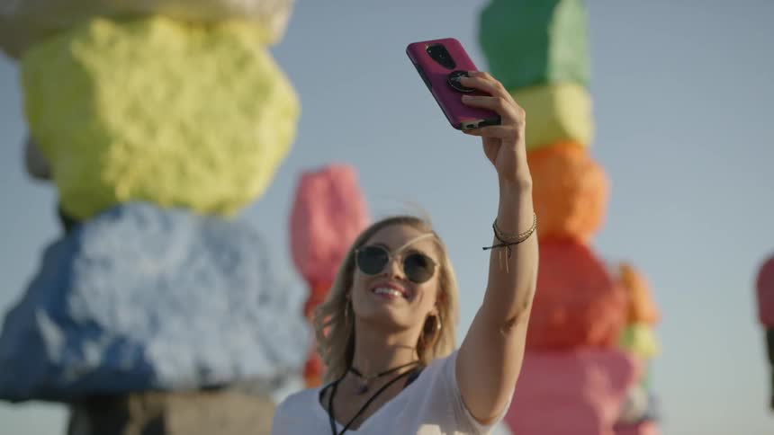 Blonde woman wearing sunglasses takes a selfie in front of the colorful stone pillars outdoors
