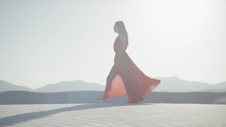 Brunette woman wearing a red dress walks along the the sand desert while the sun shines