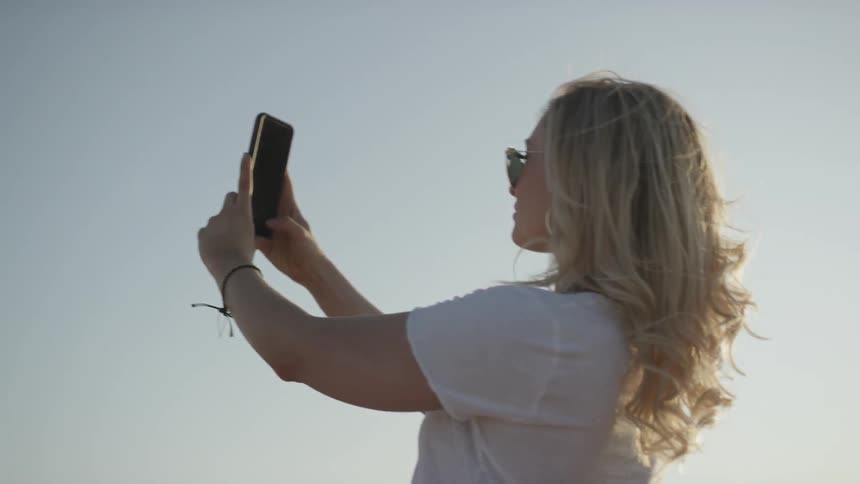 A blonde haired woman wearing sunglasses takes photos at low sun