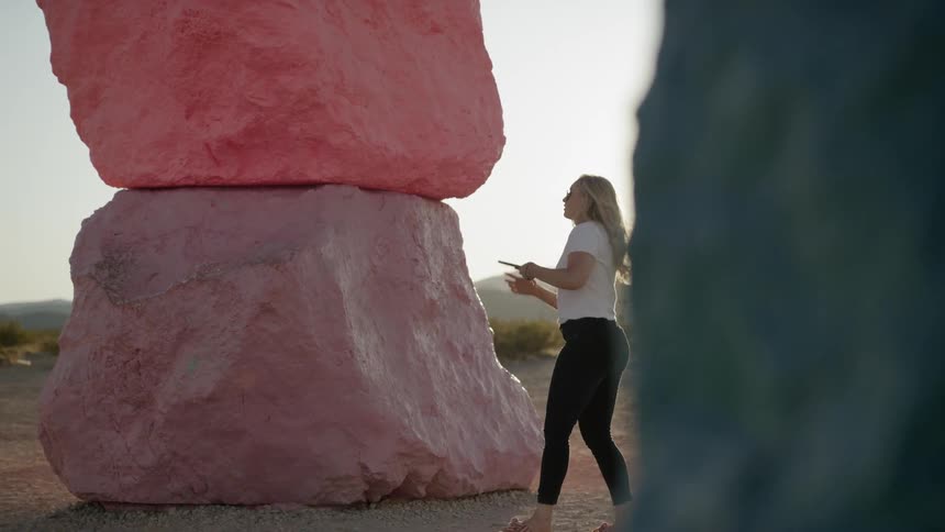 Girl walking and touching a large rock before pulling out cell phone