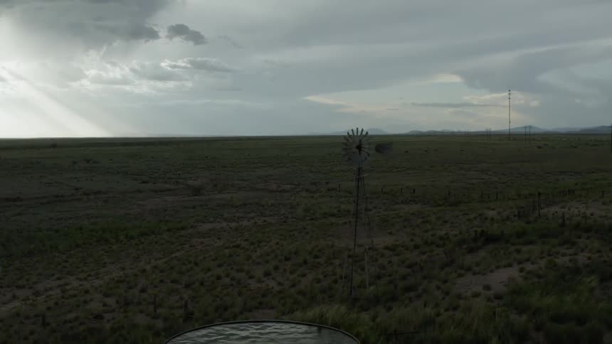 A windmill stands beside a water tank on a brush filled landscape on a cloudy day