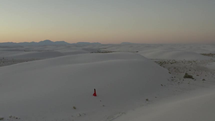 Brunette woman wearing a red dress walks along the white sand desert at sunset