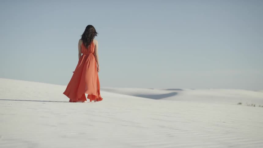A woman in a full length dress walking on sand dunes looking out to the horizon