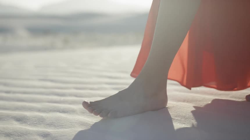 A womans feet with a long orange dress walking on the desert sand at daytime