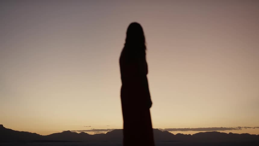 A woman in a full length dress looks out to hillside on the horizon at sunset