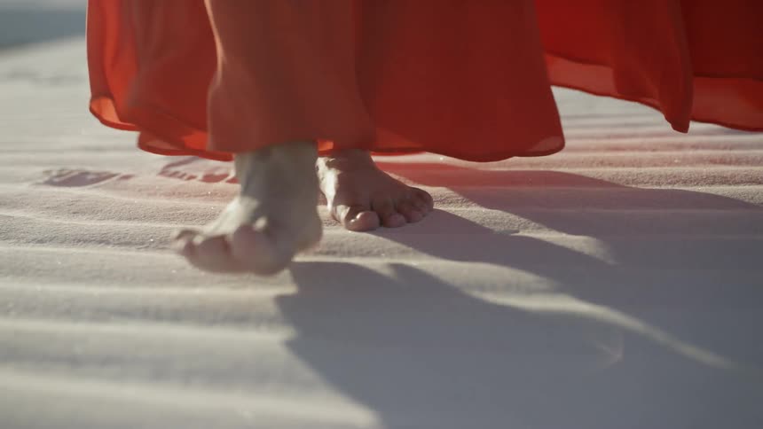 A woman in a long billowing dress walks barefoot over sand in a desert