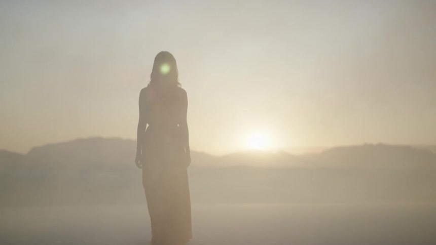A woman in a red gown walks on the desert sand as wind blows and the sun sets