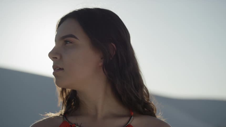 Brunette woman wearing a red dress looks sideways in the desert while the sun shines