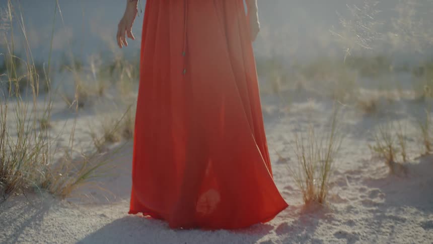 A barefoot woman in a red gown walking through the sand in a desert during the day