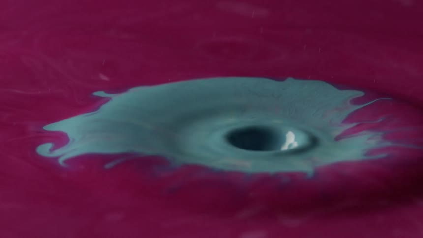 A pool of purple paint with a blue blotch in the middle ripples as a drop of blue paint falls on the blue blotch