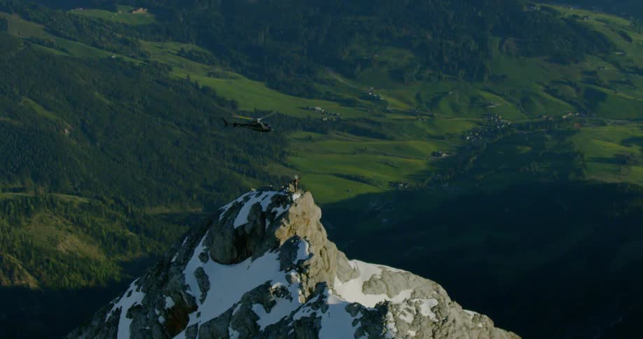 Helicopter flies over snow cover peak with forested mountain occupying background