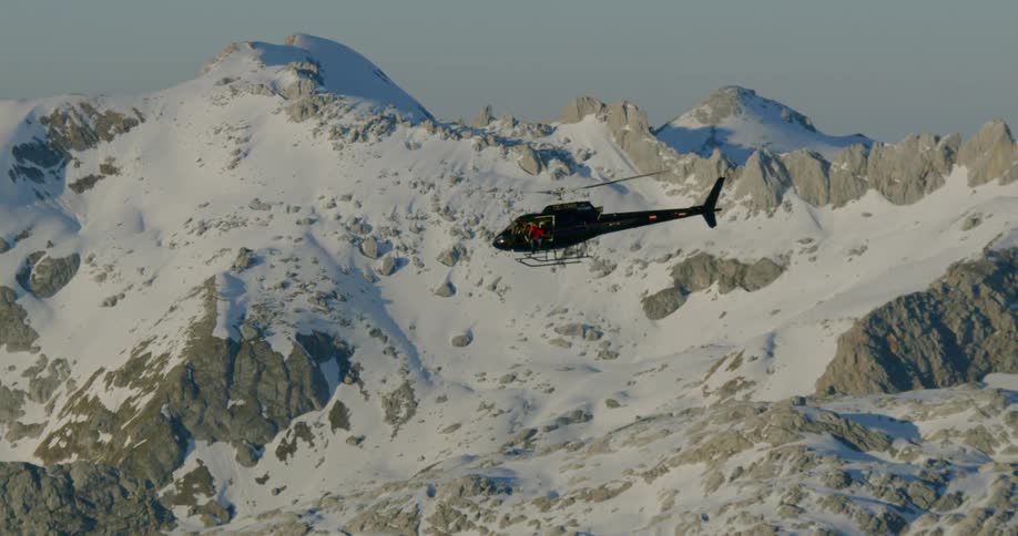 Man sits in open door of helicopter flying over snowy mountain range