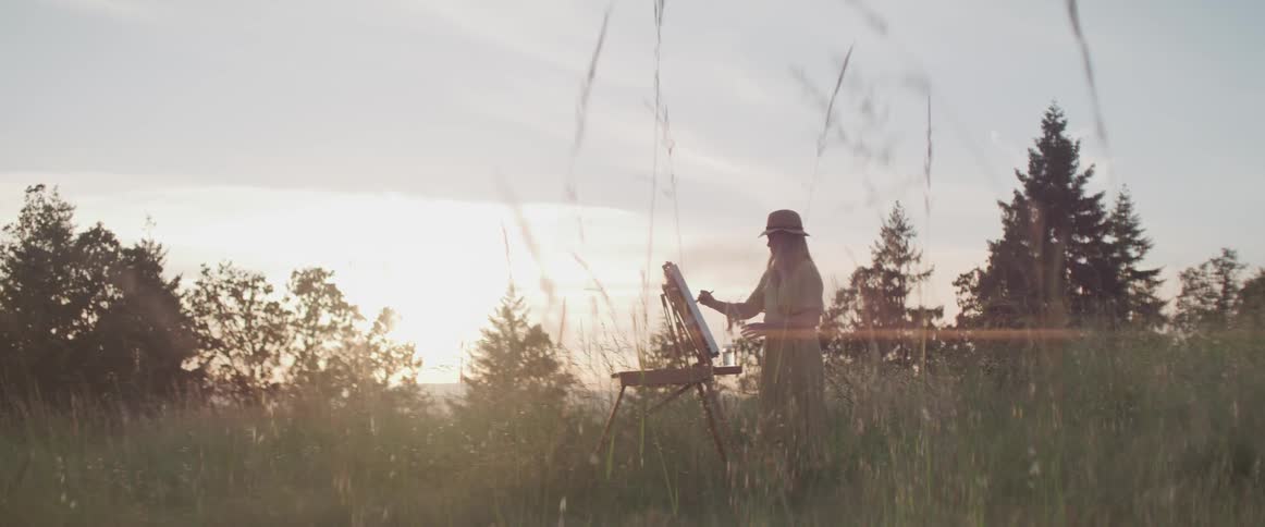 A woman in a hat paints at sunset