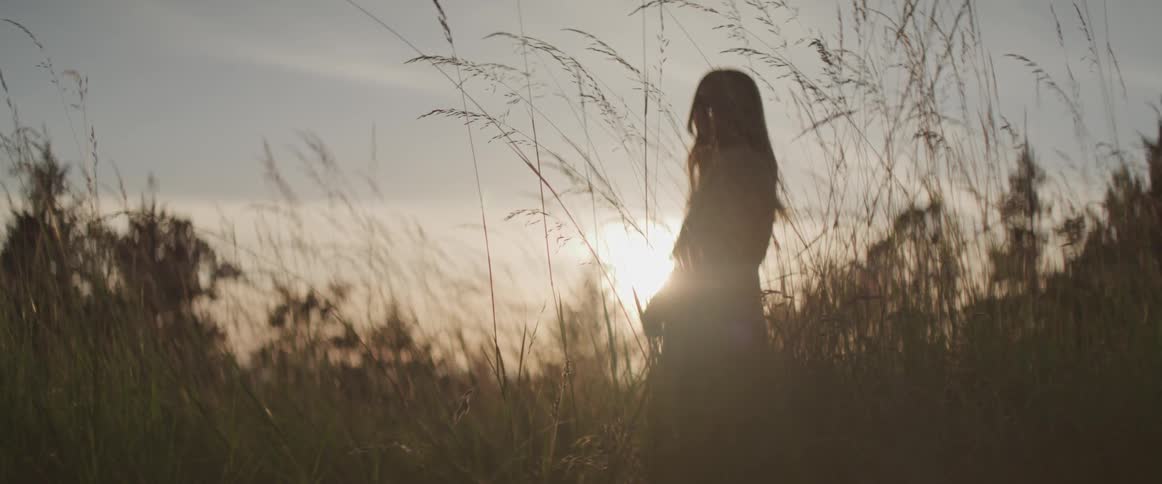 A woman stands in a meadow with the sun bright behind her