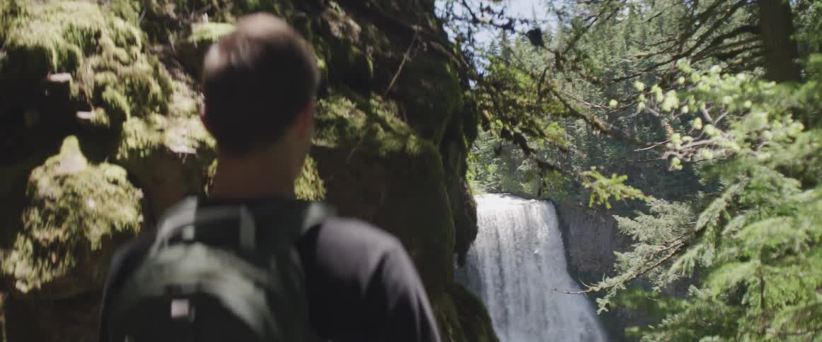 Man wearing a backpack looks at the waterfall in the green woods