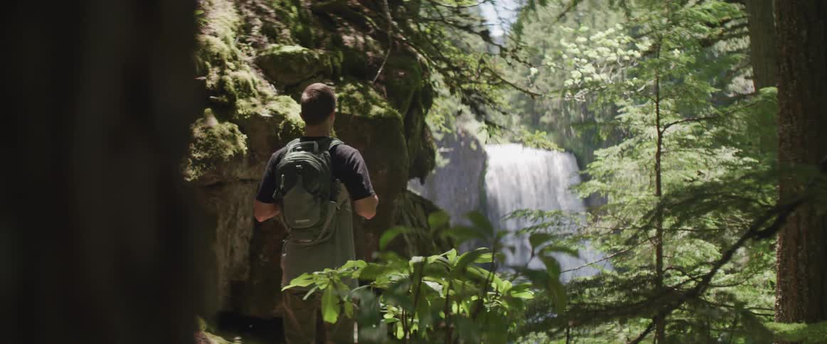 A man stands on the edge of a cliff looking to the woods and waterfall