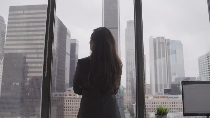 A woman stands in an office facing the floor to ceiling windows looking out at the city