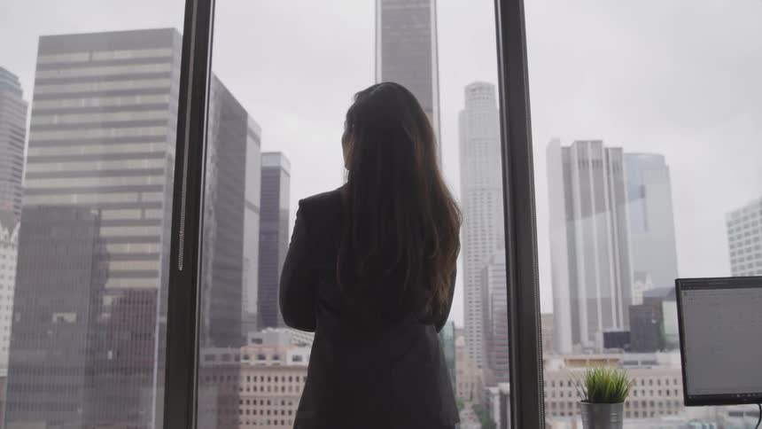 Woman standing with arms crossed looking out an office window to the city