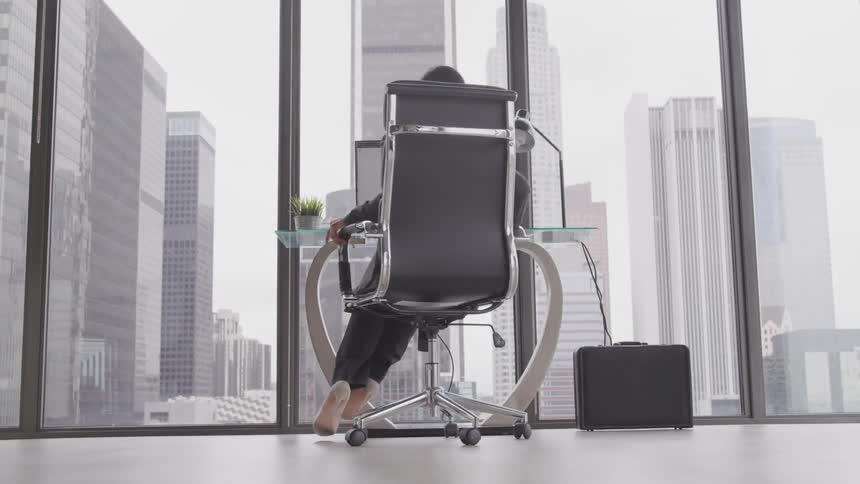 A woman walks in a room and sits in a chair at her desk