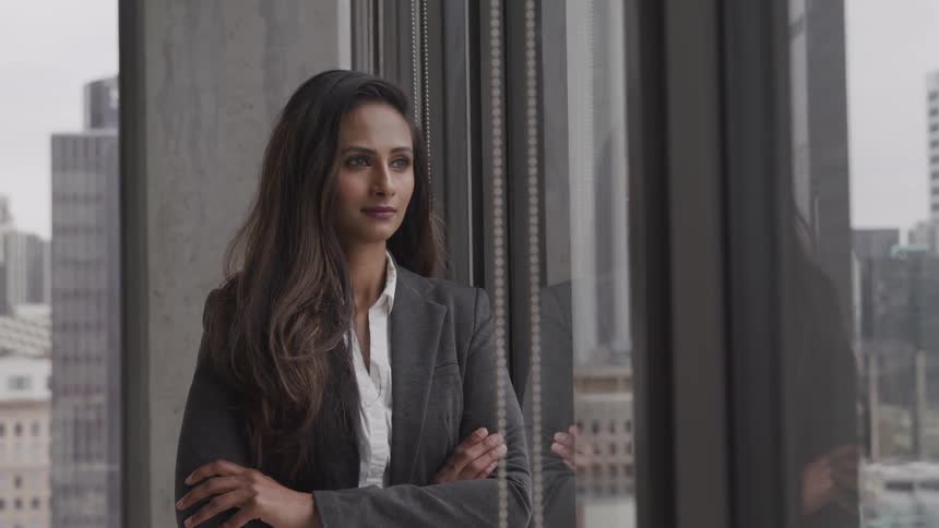 A woman stands in an office building beside a large set of windows looking outside to the city