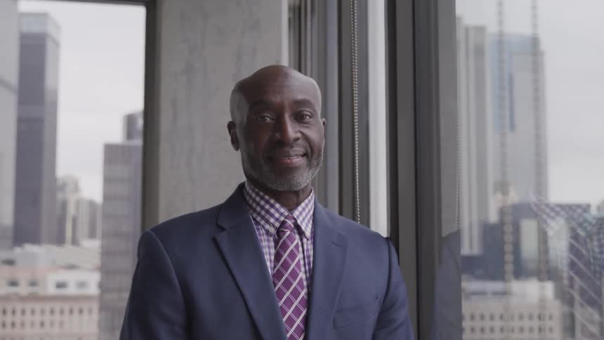 Bald headed businessman stands next to the window and looks at the camera smilingly in the office