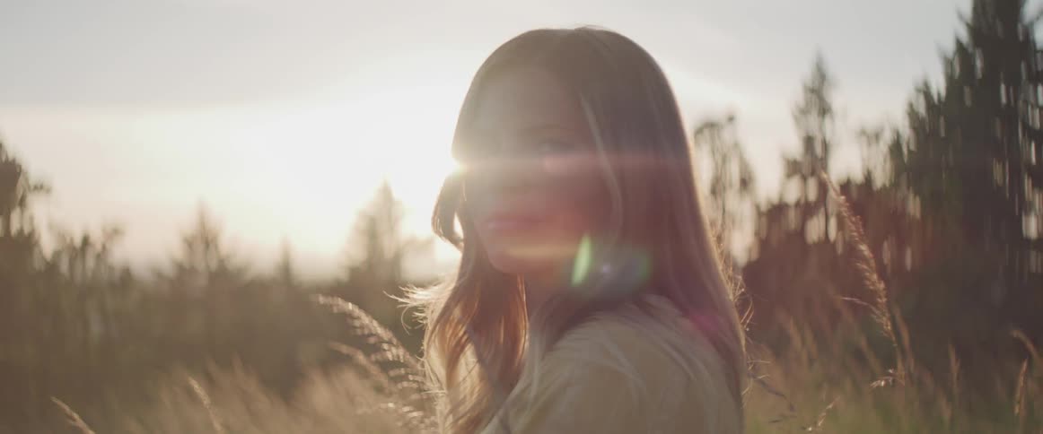 Blonde woman standing in the tall dry grass looks at the camera at sunset