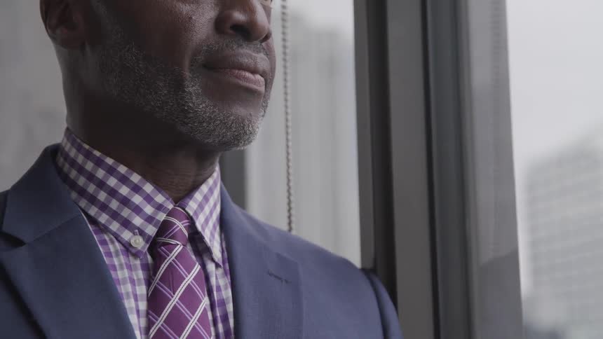 Black bald bearded businessman wearing a suit stands next to the window in the office
